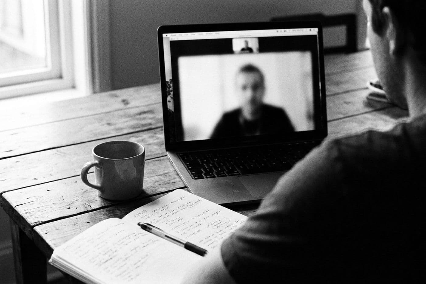 A video call on a laptop at a timber table, a notebook of handwritten notes beside it.