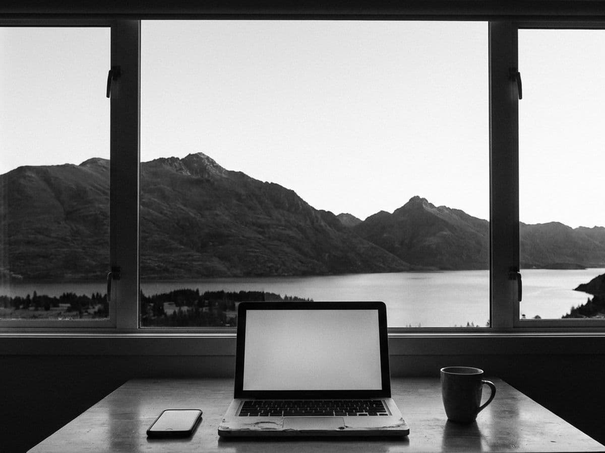 A laptop on a desk in front of a window looking out at mountains and a lake.