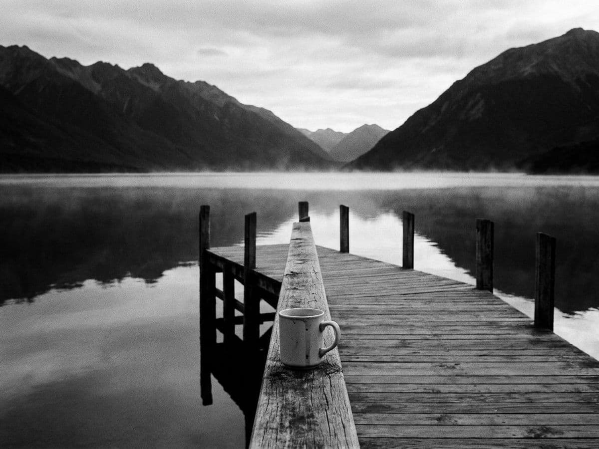 A coffee mug on a jetty over a still lake, mountains behind.